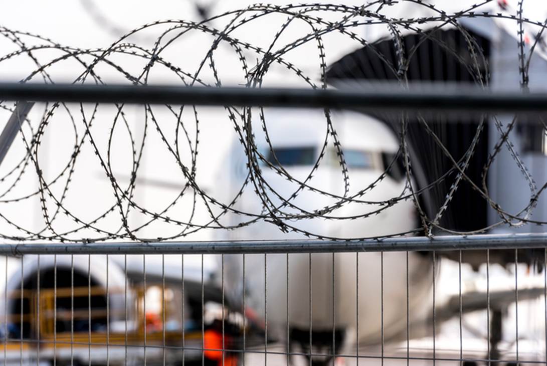 A razor wire fence is in front of an airplane at a terminal. The high security image relates to passengers' rights, even in high-security airport environments.